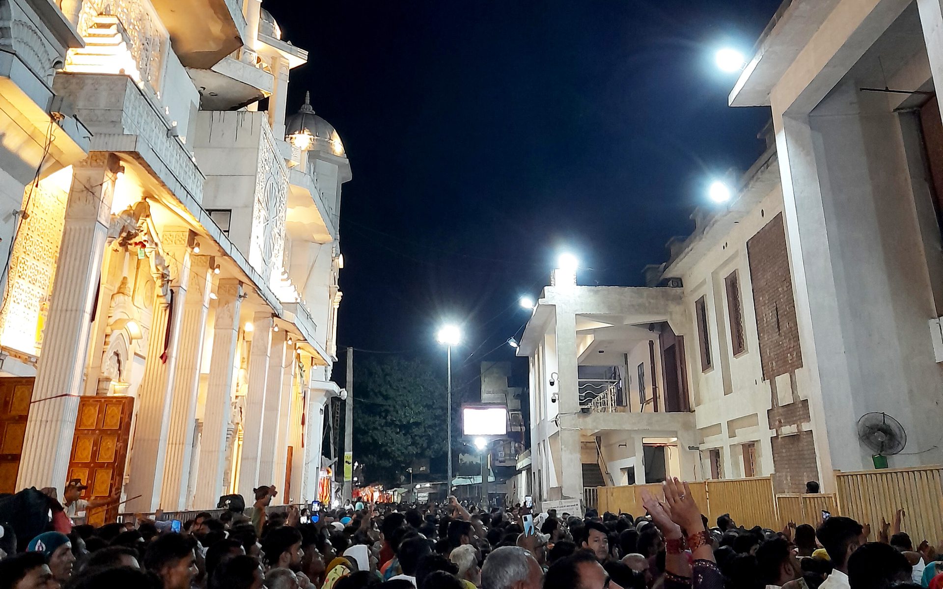 A night crowd of devotees with raised hands fills the lane in front of the floodlit white facade of Mehandipur Balaji Temple, with its domed chhatris